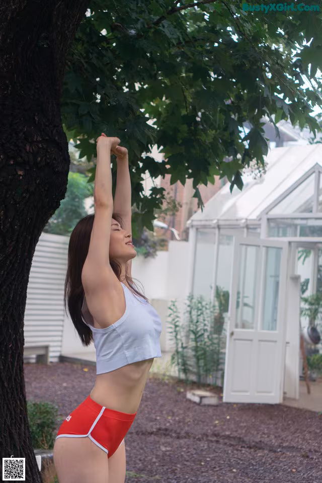 A woman in a white top and red shorts standing under a tree.