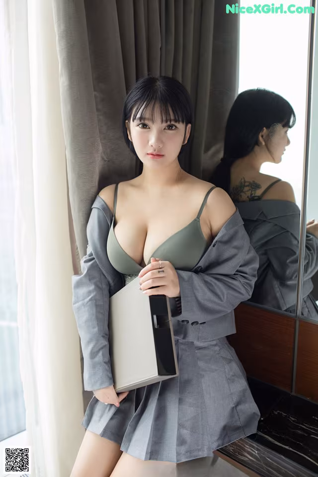 A woman in a gray dress holding a box in front of a mirror.