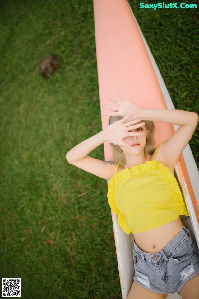 A woman in a yellow top and denim shorts leaning against a surfboard.